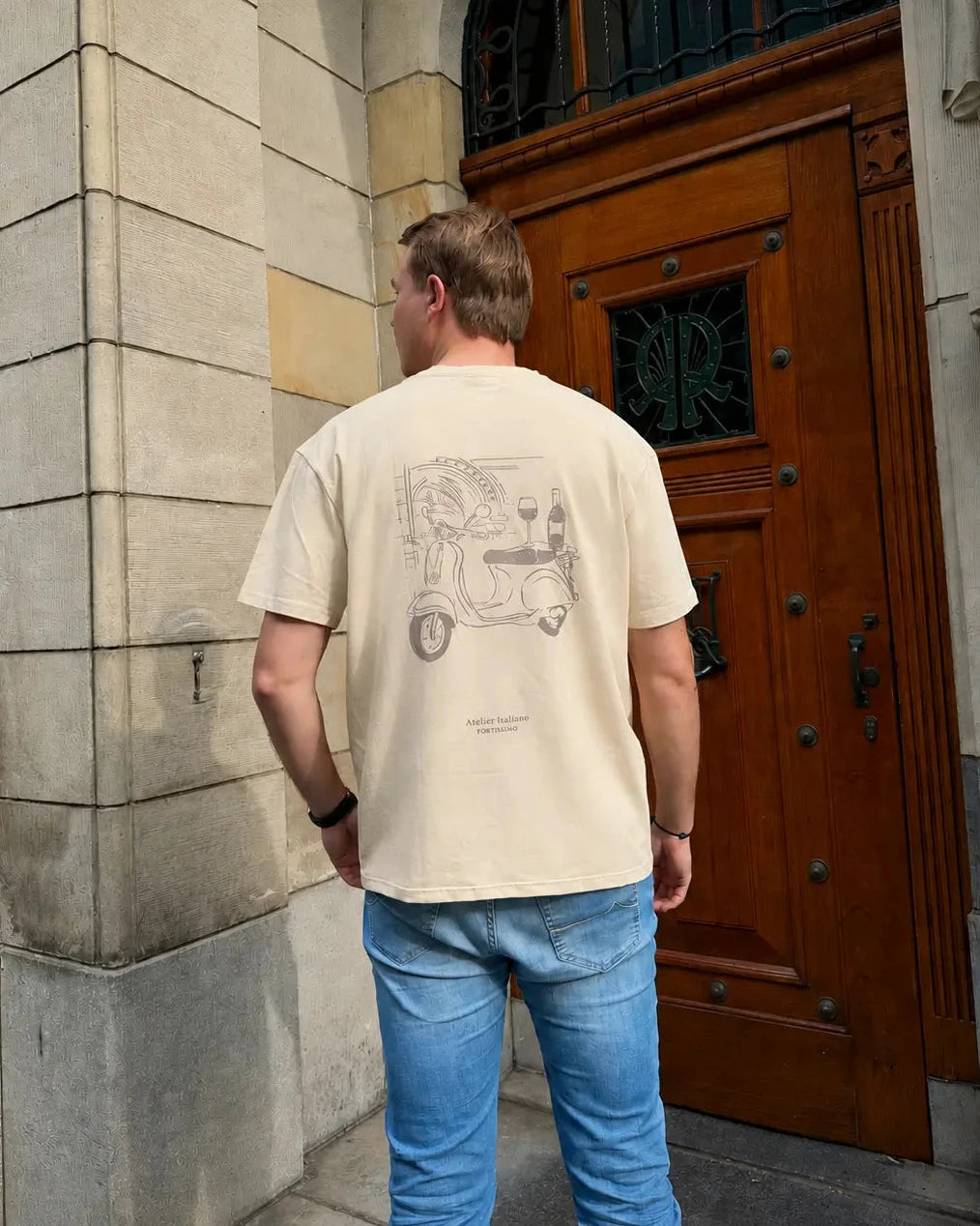 Person wearing a beige t-shirt with a graphic design, standing in front of a wooden door.