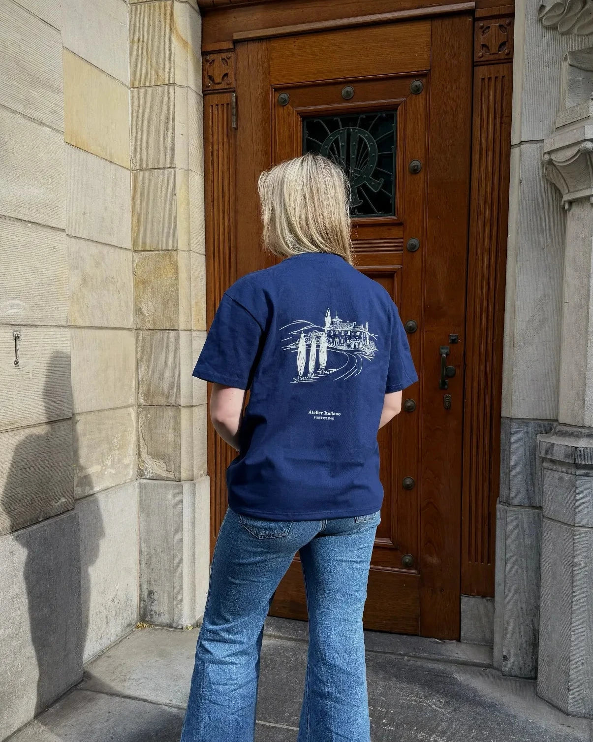 Person wearing a blue t-shirt with a logo standing in front of a large wooden door.