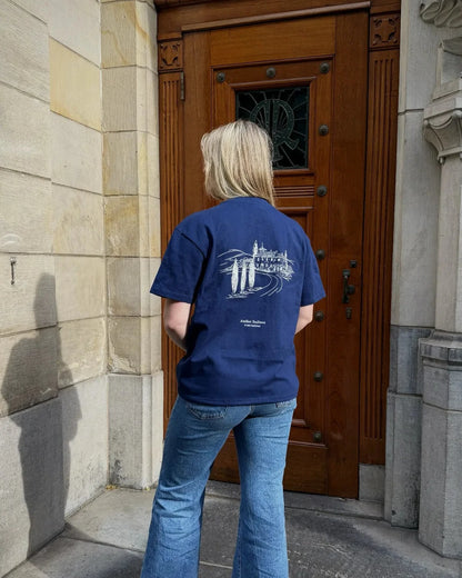 Person wearing a blue t-shirt with a logo standing in front of a large wooden door.