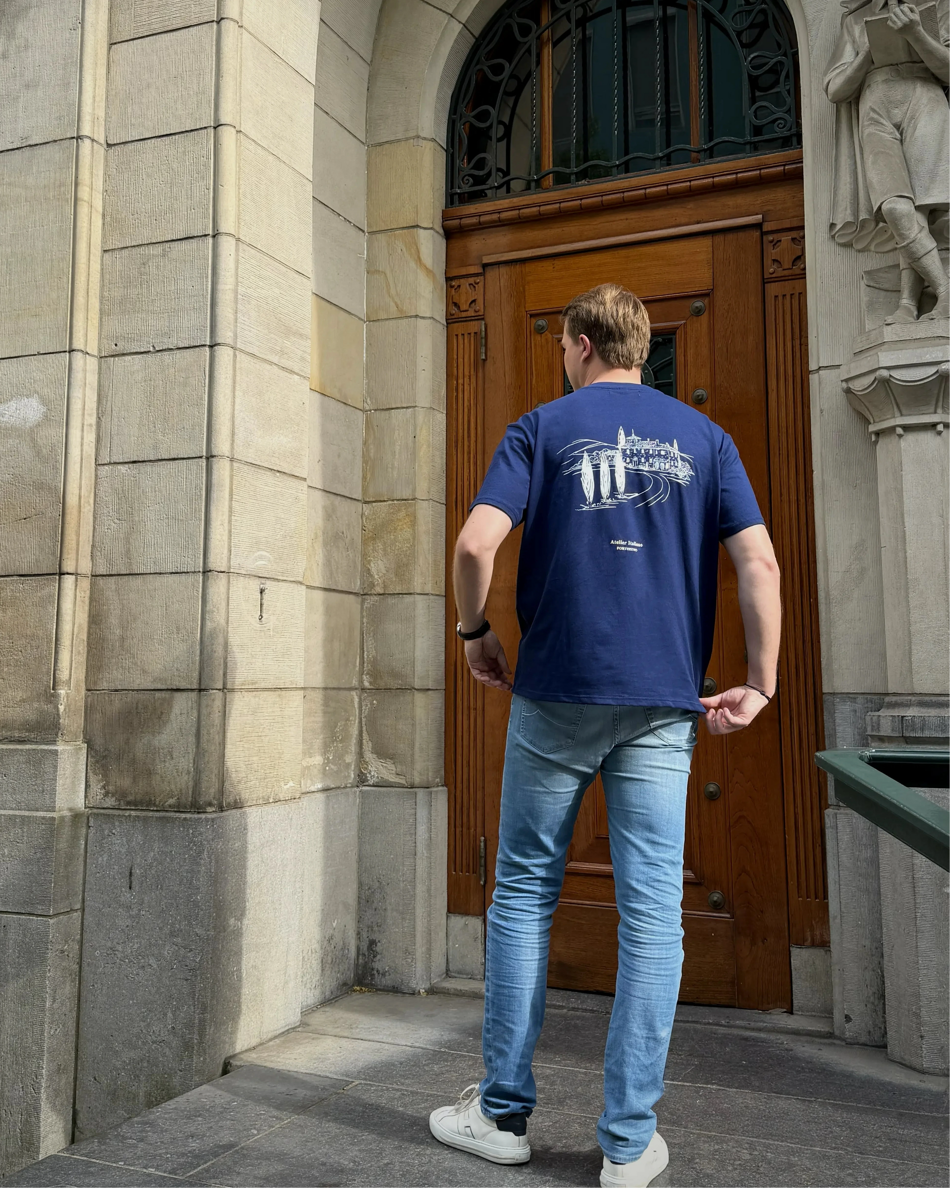 Man in a blue t-shirt and jeans standing in front of a large stone building with a wooden door.