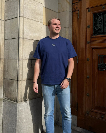 Man wearing a navy blue t-shirt and jeans standing in front of a stone wall and wooden door.